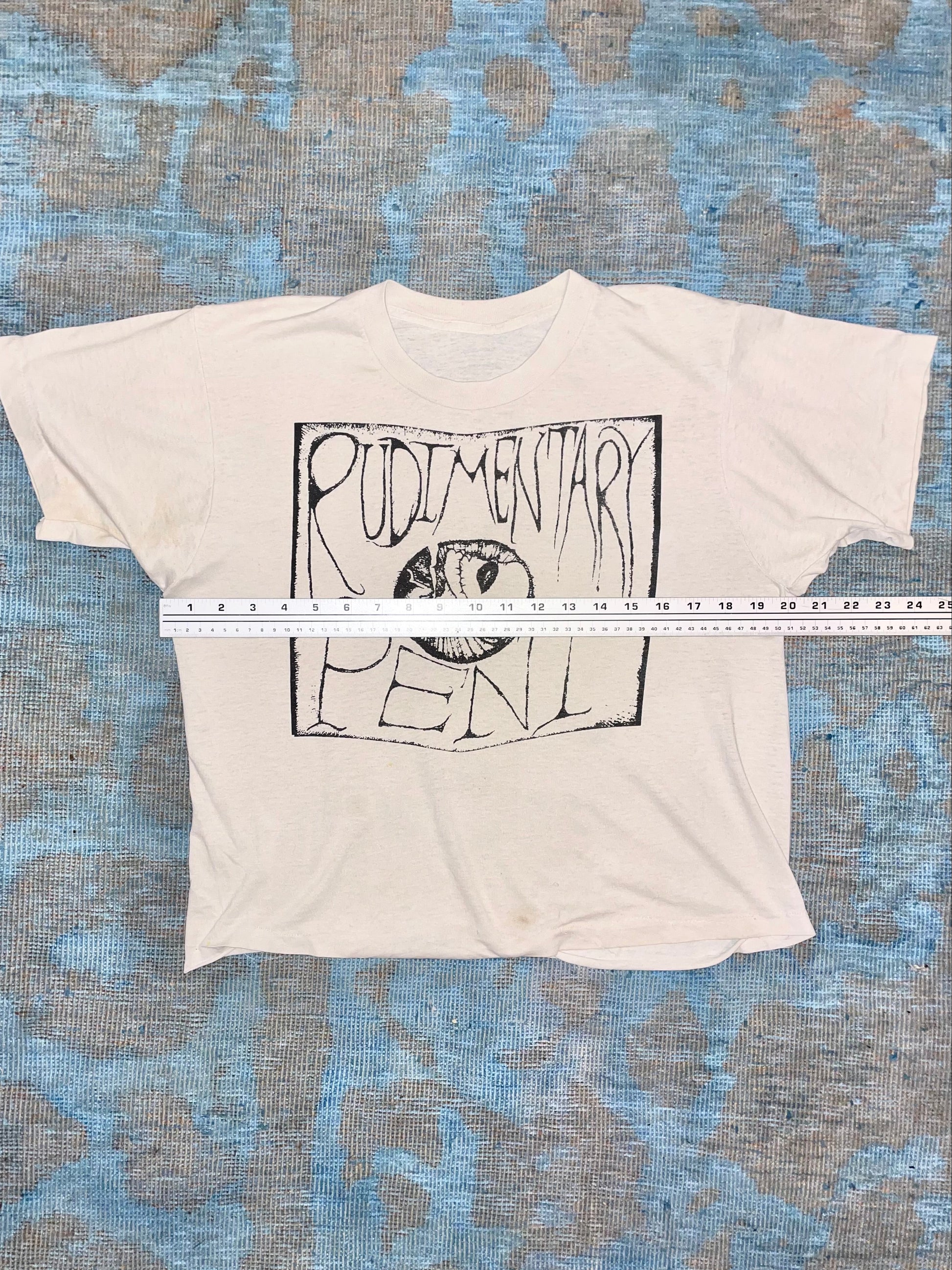 White t-shirt with black "Rudimentary Peni" graphic design on a blue textured surface